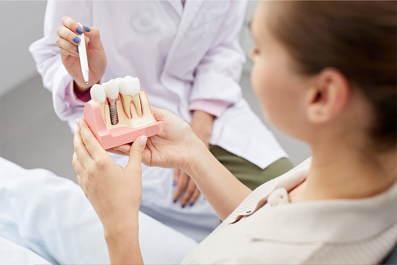 A dental hygienist demonstrates oral care techniques to a woman in a clinical setting, with the dental hygienist holding a model mouth and a toothbrush.