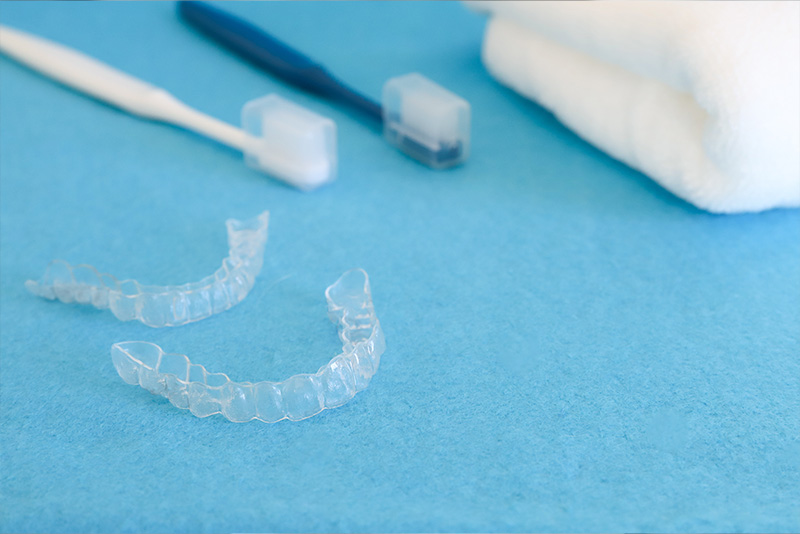 The image shows a blue toothbrush and two sets of clear dental braces, with one set having the upper braces in place and the lower braces removed, lying on a light-colored surface.