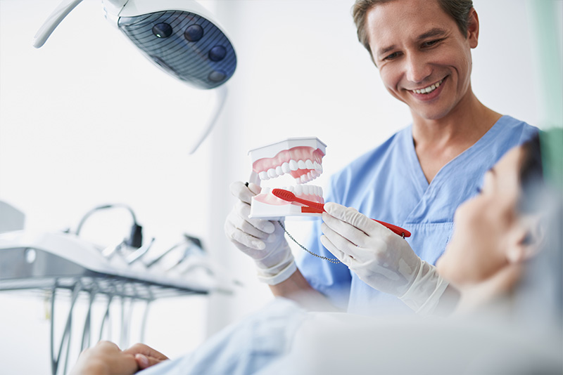 A dental professional in a white lab coat and surgical mask, holding a red toothbrush, smiling at the camera while standing behind a patient receiving care.
