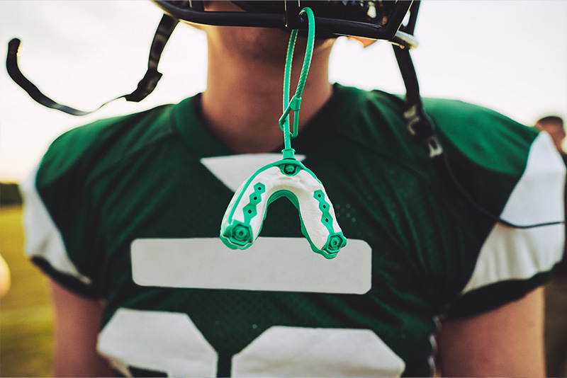The image shows a football player wearing a helmet with a green and white color scheme, holding what appears to be a mouthguard. He is dressed in a uniform that includes a jersey with the number 56 on it, along with other team-related insignia. The player s gaze is directed off-camera, and he seems to be looking into the distance. The background is blurred but suggests an outdoor setting, possibly a field or stadium. There are no visible texts or distinguishing marks that provide additional context about the player or the event.