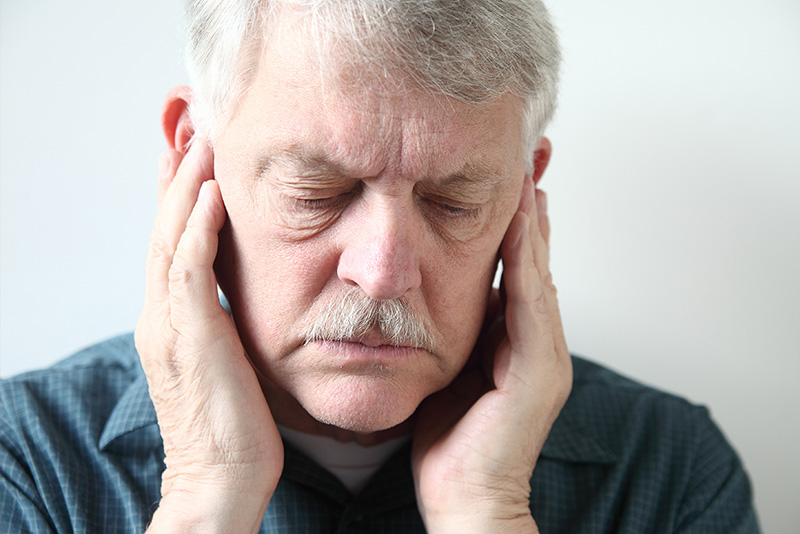 The image shows an elderly man with a beard, wearing glasses and a dark shirt, holding his hand to his ear in a thoughtful or contemplative pose.