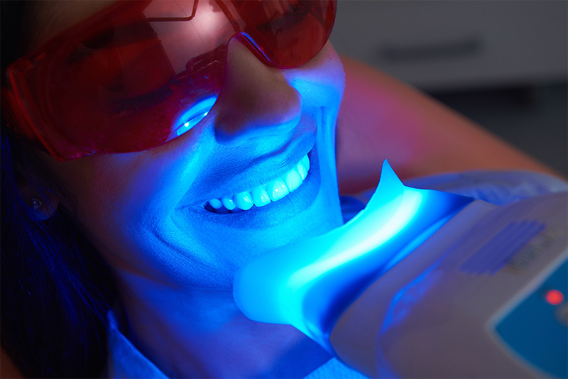A woman with a blue light around her neck, possibly from a dental device, is smiling at the camera.