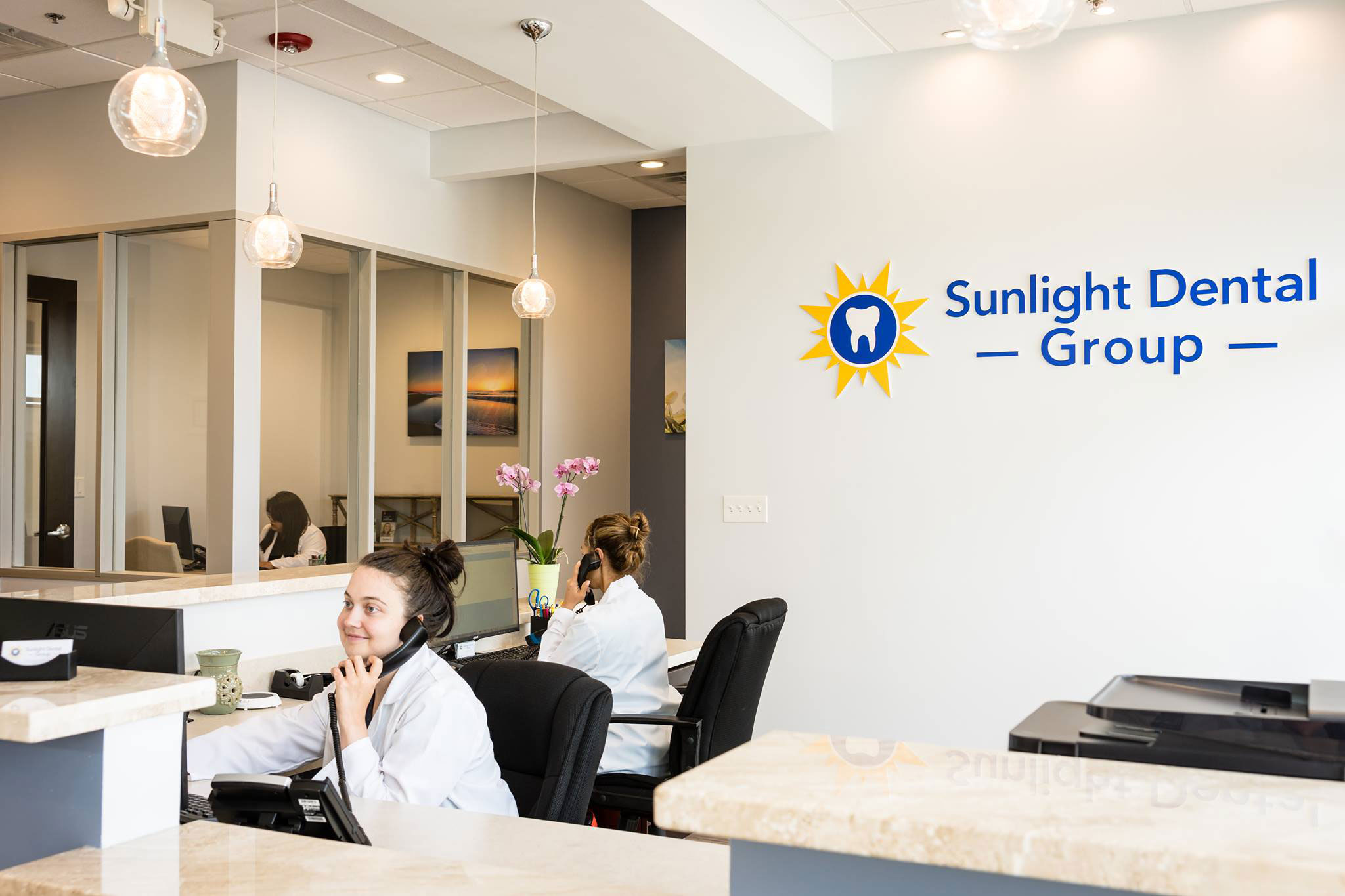 The image shows an interior view of a dental office, featuring a reception area with a woman sitting at the desk and a sign that reads 'Sunlight Dental Group.