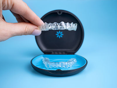 The image shows a hand holding an open box containing a set of clear, plastic aligners, which are used for teeth straightening.