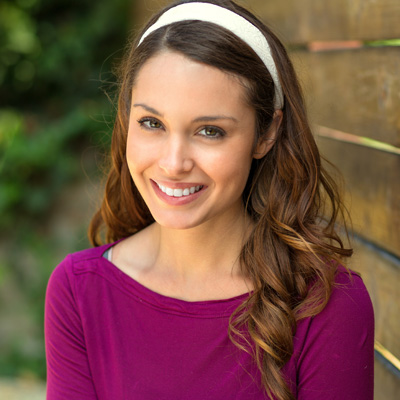 The image shows a woman with long hair smiling at the camera, wearing a purple top and a headband. She is standing against a wooden fence or wall.