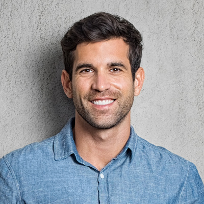A bearded man with a friendly smile, wearing a blue shirt, poses against a concrete wall.