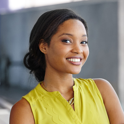 Alt text   A smiling woman with dark hair, wearing a yellow top and posing against a blurred background.