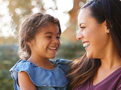 A woman and a child are smiling at the camera, with the woman holding the child. They appear to be outdoors during the daytime.