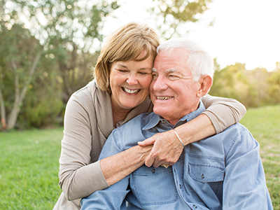 The image features an elderly couple, a man and a woman, embracing each other in a warm hug. They are outdoors on what appears to be a sunny day, with the man wearing a blue shirt and the woman in a light-colored top.