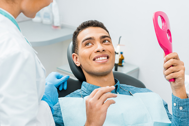 A man sitting in a dental chair, smiling broadly, with a dental hygienist standing behind him holding a red toothbrush.