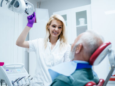 A woman in a white lab coat stands beside an elderly man in a dentist s chair, holding a dental mirror.