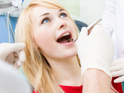 The image shows a woman seated in a dental chair, receiving dental care from a professional. She is smiling and appears to be in the process of having her teeth cleaned or checked.