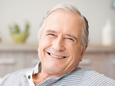 The image shows a smiling elderly man with gray hair, wearing a dark shirt and sitting in a relaxed posture.