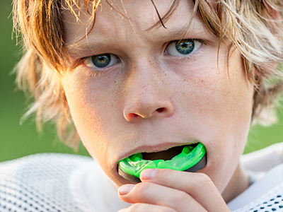 The image shows a young boy with blonde hair, wearing a football uniform and holding a green object to his mouth. He appears to be in the midst of an activity or sport, as suggested by the uniform and the context provided by the object he s holding.